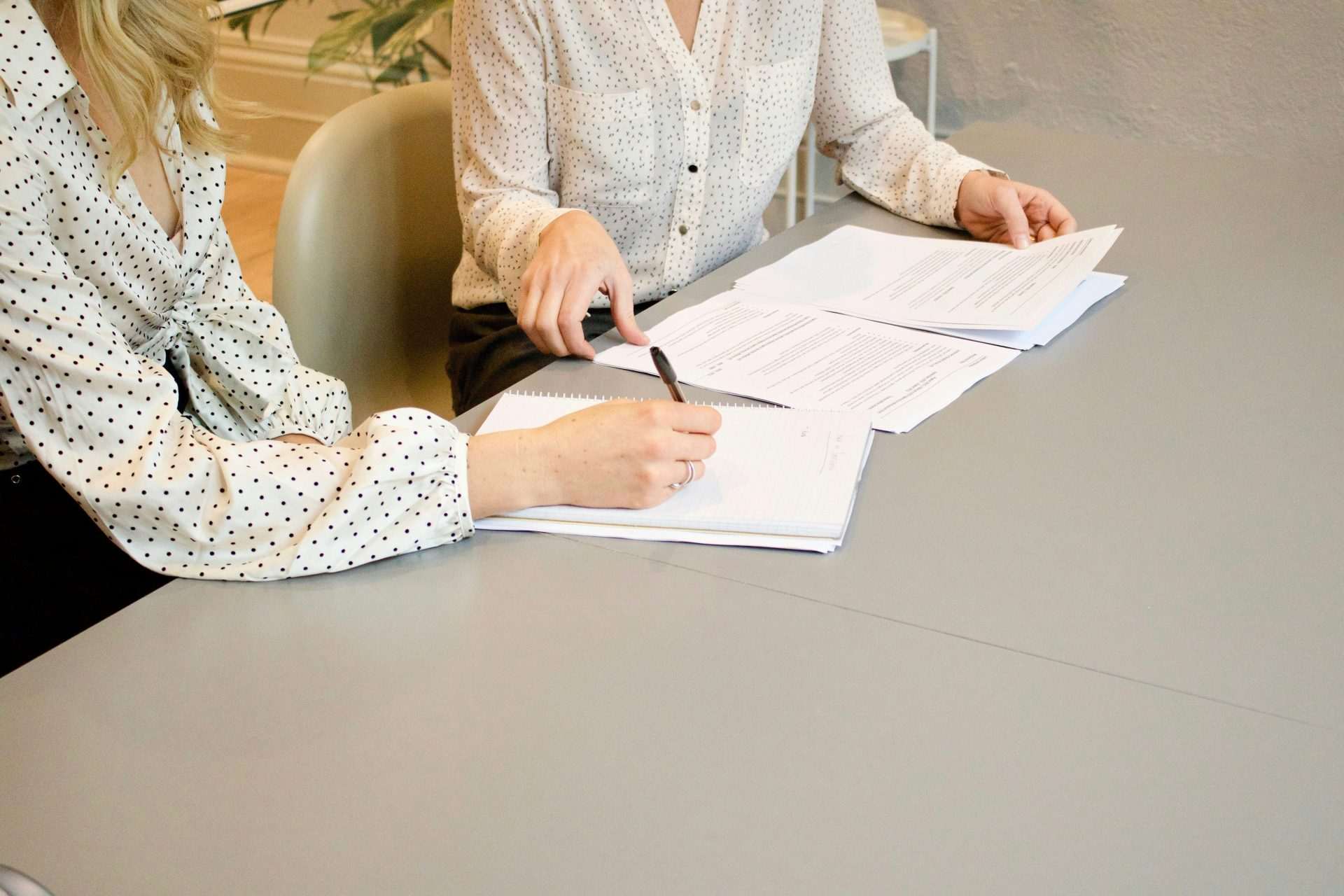 Une femme signe sur une feuille de papier blanc d'imprimante à côté d'une autre femme qui s'apprête à toucher les documents